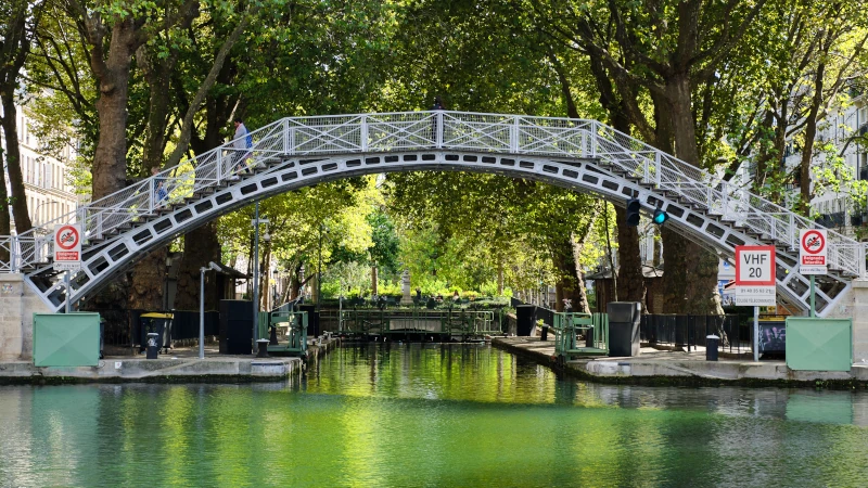 Iconic footbridge of the Canal Saint-Martin in Paris, with people crossing and lush green trees in the background.