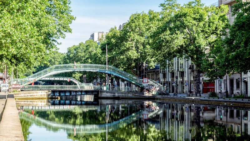 Footbridge over the Canal Saint-Martin in Paris, surrounded by trees and buildings, with a perfect reflection in the calm water.