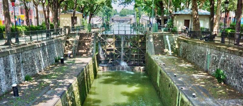 Écluse du Canal Saint-Martin à Paris, entourée de végétation et de chemins piétonniers ombragés, avec des chutes d'eau aux murs latéraux.